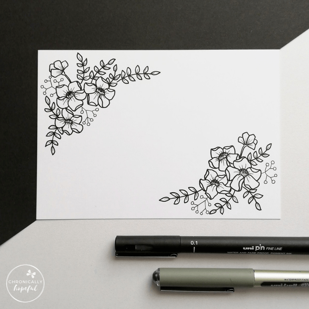 A white card with black botanical drawing, two pens positioned below the card on the table.
