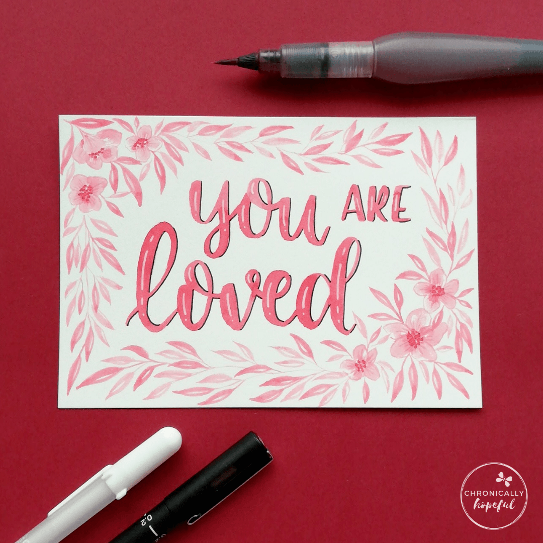 You Are Loved, watercolour lettering with botanical frame, pink paint on white card, Paint brush and pens on the table around the card.