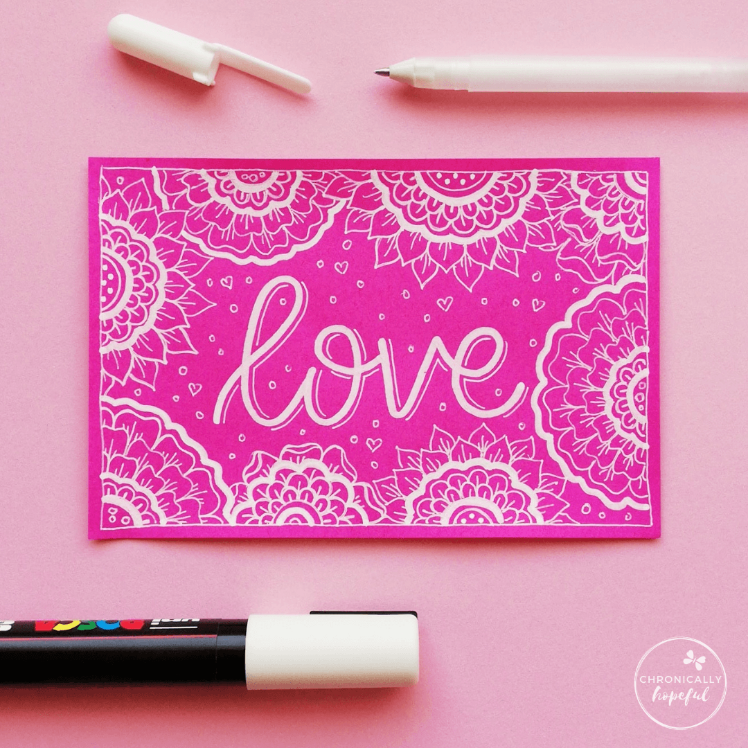 Pink card with the word Love lettered in white paint pen with floral mandalas drawn around it. 2 pens positioned on the table around the card.