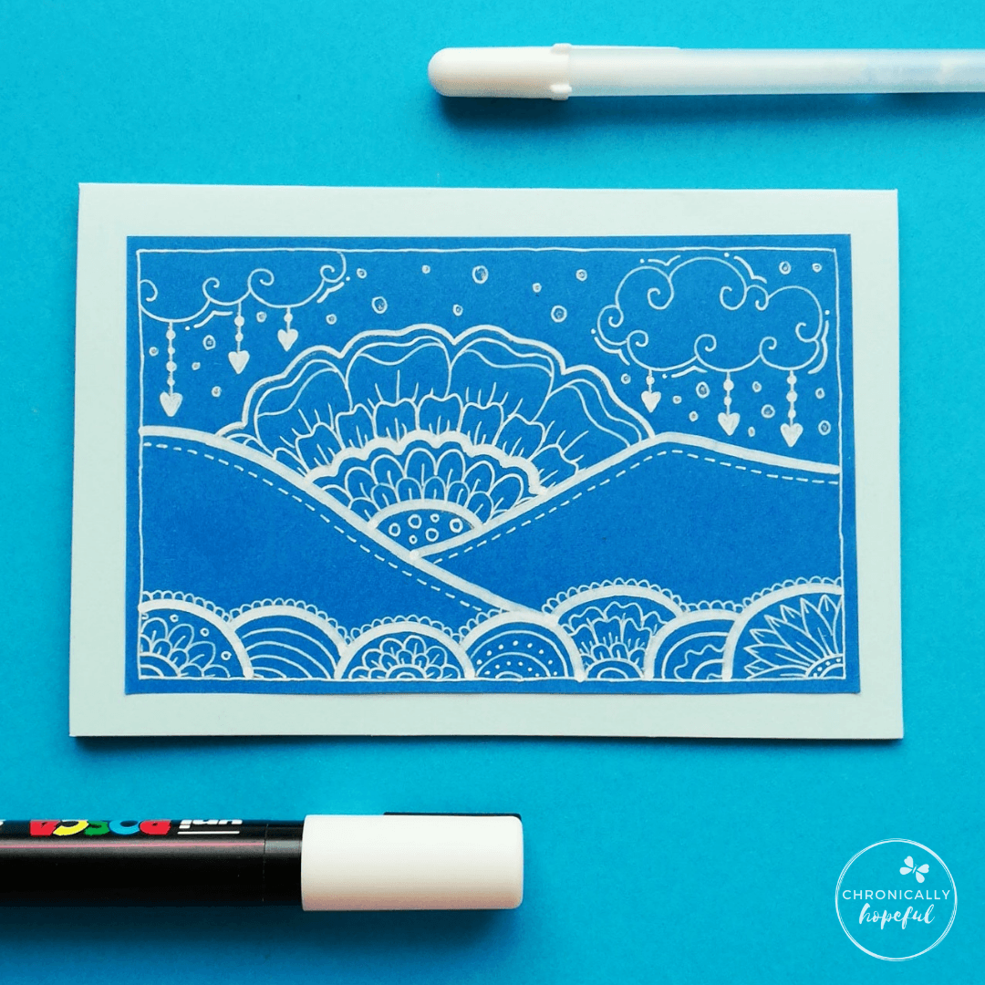 Whimsical scene of hills, sunset and clouds drawn in white paint pen on blue card. 2 pens on the table around the card.