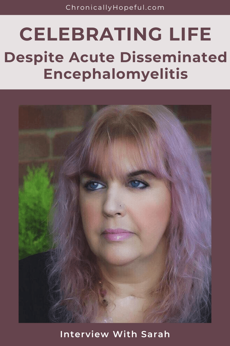 Sarah standing in front of a brick wall. She has pink hair and a black top. Title reads: Celebrating life despite Acute Dissemminated Encephalomyelitis. Interview with Sarah.