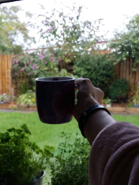 holding up a mug of coffee in front of a window overlooking a very green garden
