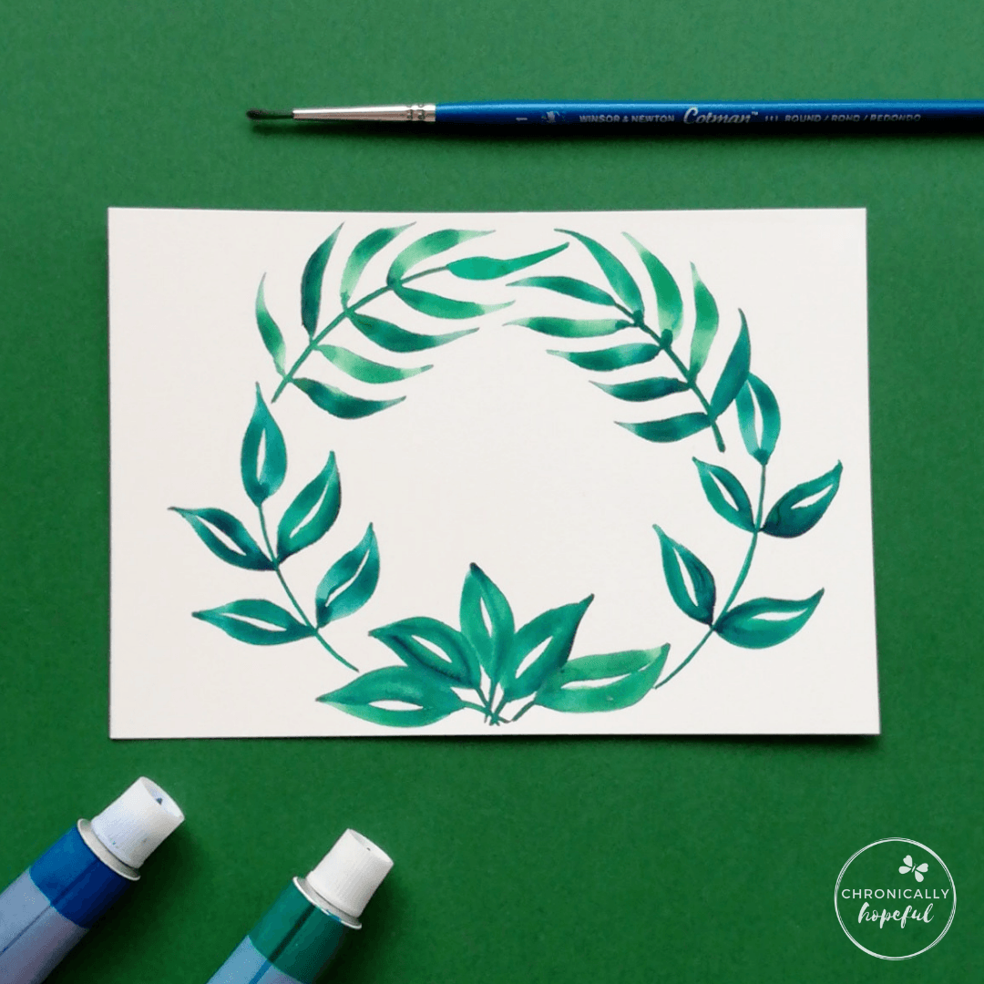 Leafy green watercolour wreath on white card, paint tubes and brush on table around the card