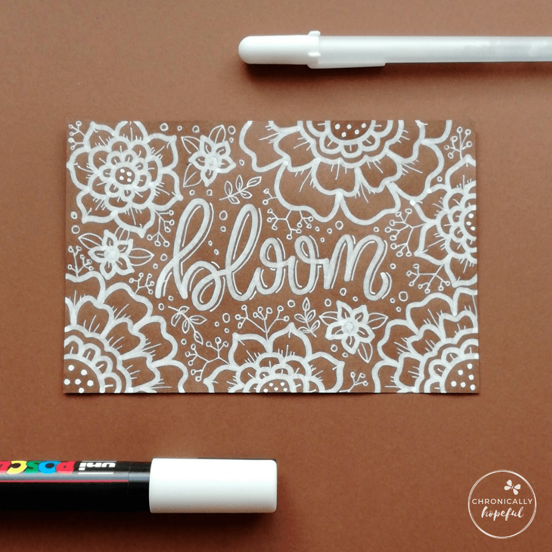 A brown card with the word Bloom lettered in cursive with white paint pen. 2 pens positioned around the card on the table.