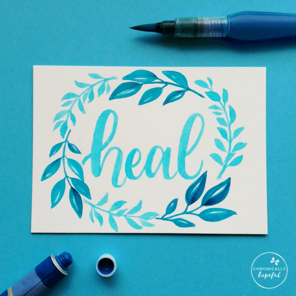 The word heal lettered in blue watercolour framed by a blue leafy wreath painted on white card by Chronically Hopeful Char