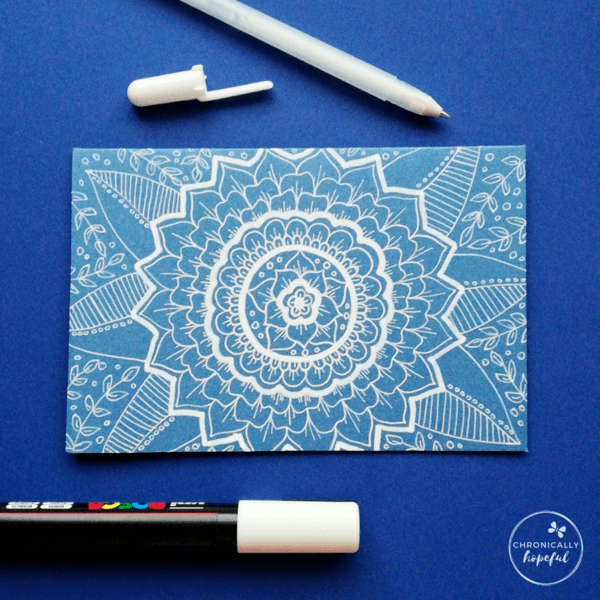 Floral mandalas in white paint pen on blue card, 2 pens on the table around the card.