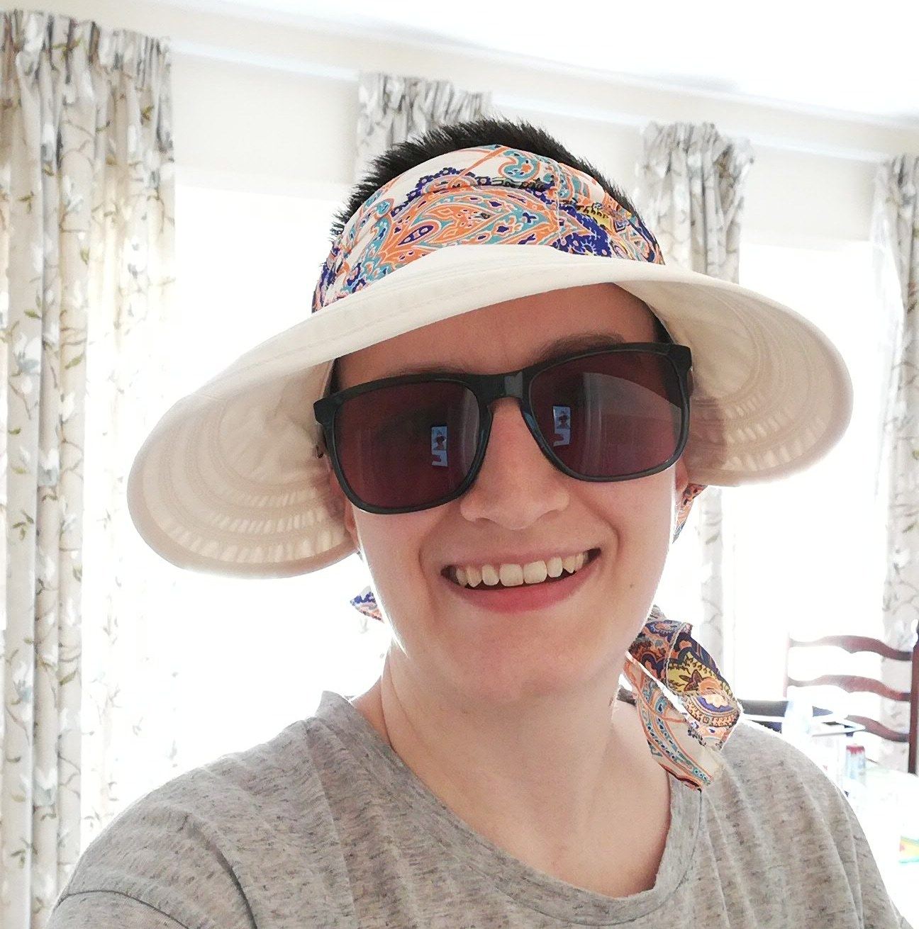 Char wearing a wide-rimmed summer hat and sunglasses. There are large windows behind her with floral curtains hanging.