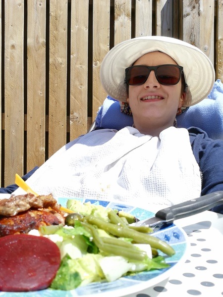 Char lying on garden recliner with a plate of food. She's wearing a sun hat, sunglasses and a bib.